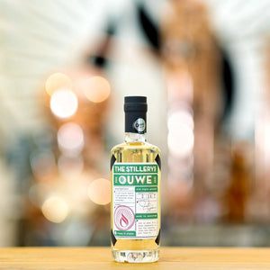 The Stillery's Ouwe Genever: bottle on a wooden bar, copper stills blurred in the background