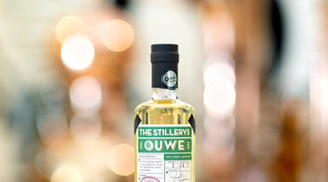 The Stillery's Ouwe Genever: bottle on a wooden bar, copper stills blurred in the background